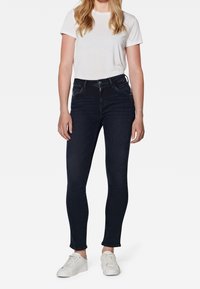 Jeans skinny biru tua terbuat dari denim, dilengkapi dengan saku depan dan ritsleting, dipadukan dengan kaos lengan pendek berwarna putih dengan leher bulat dan sepatu sneakers putih.