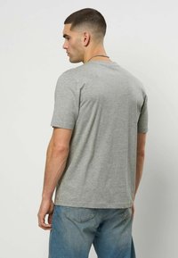 Mann med kort hår iført en grå kortermet t-skjorte og blå jeans, stående med ryggen mot en ensfarget lys bakgrunn.