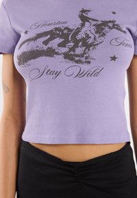 Purple ribbed cropped top featuring a black cowboy graphic, text "Houston Texas" and "Stay Wild," accented with small stars. Black waistband below.