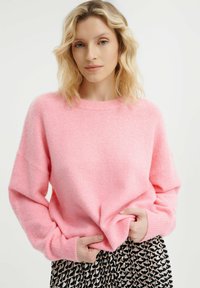 Roze oversized gebreide trui met een ronde halslijn en geribbelde mouwen, gecombineerd met een zwart-wit gestreepte rok. Zachte textuur is zichtbaar.