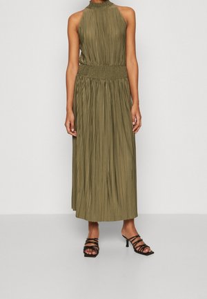 Długa sukienka maxi w kolorze oliwkowej zieleni z pionowymi plisami, z wiązaniem na szyi i marszczonym paskiem w talii. Noszona z czarnymi sandałami na wysokim obcasie z paskami.