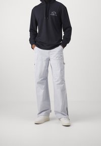 Svart huvtröja med logotyp, tillsammans med ljusgrå cargobyxor med sidofickor. Vita sneakers fulländar outfiten.