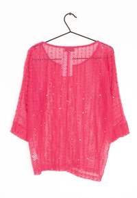 Haut rose en maille avec une coupe ample, des manches chauve-souris, un col rond et des sequins tissés dans le tissu. Accroché à un cintre noir en fil.