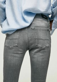 Jeans de denim gris con un corte slim, que cuentan con dos bolsillos traseros y un diseño cosido. Combinados con una camisa de rayas azul claro.