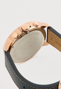 Ροζ χρυσή πίσω πλευρά ρολογιού με χαραγμένο το λογότυπο της μάρκας, δύο πλευρικούς διακόπτες και σκούρα δερμάτινη ταινία με ορατή ραφή.