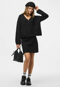 Zwarte cropped sweater met een rits en twee voorkruiszakken, gecombineerd met een zwarte minirok. Accessoires zijn een zwarte gevlochten handtas en gestreepte sokken.