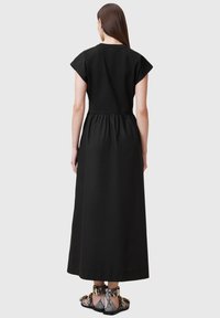 Vestido maxi negro con mangas cortas, cintura fruncida y un corte holgado. La modelo lleva botas de tobillo texturizadas con un patrón de serpiente.