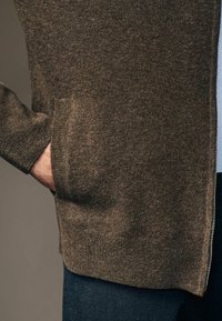 Brązowy dzianinowy kardigan o strukturalnym wykończeniu, z boczną kieszenią i zamkiem błyskawicznym wzdłuż krawędzi. Styl casualowy, miękki materiał.