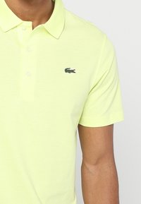 Lime zelené polo triko s límečkem, obsahující tříknoflíkovou légu a vyšívané logo krokodýla na hrudi.