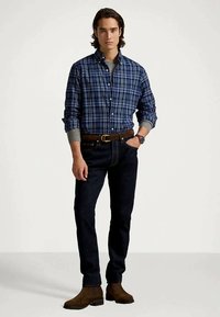 Man med blå rutiga skjorta över grå tröja, mörka jeans, brun bälte, klocka och bruna stövlar, står med en hand i fickan.