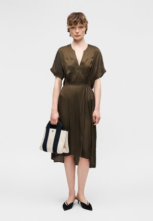 Γυναίκα που φοράει μίντι φόρεμα σε ελιά πράσινο χρώμα, κρατά μια μπεζ και ναυτικό τσάντα, και φοράει μαύρα γοβάκια με μυτερή μύτη και μικρό τακούνι, στέκεται μπροστά σε λευκό φόντο.