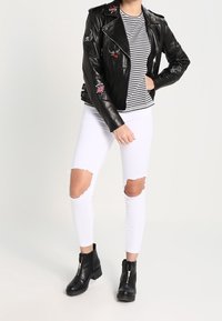 Chaqueta de motociclista de cuero negra con parches bordados, camiseta a rayas, jeans blancos rasgados y botas negras al tobillo con cierre de cremallera.