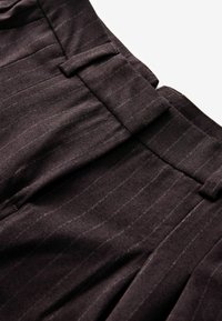 Tumed pruunid pükstega, millel on peened triibud, volditud eesmine osa ja küljetaskud. Kude näib olevat tekstuurne ja struktureeritud sobivuse tagamiseks.