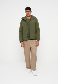 Green padded jacket with a hood, textured knit sweater underneath, beige trousers, and white sneakers. Features a small logo on the chest.