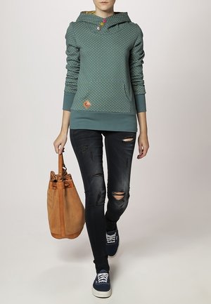 Tyrkysová mikina s puntíky a klokaní kapsou, doplněná třemi knoflíky, v kombinaci s tmavými roztrhanými skinny džíny a béžovou kabelkou přes rameno.