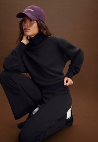 Felpa nera a girocollo con polsini a coste, abbinata a pantaloni neri a gamba larga. Un cappellino viola con la scritta "NEW YORK" arricchisce l'outfit.