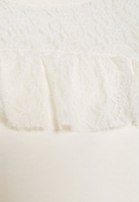 Bílá látka s horizontálním volánem z květinové krajky, který je překryt hladkým pleteným materiálem.