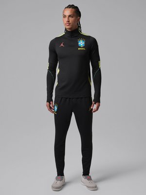 Άνδρας με πλεξούδες που φοράει μαύρο σετ προπόνησης ποδοσφαίρου της Βραζιλίας με κίτρινες λεπτομέρειες και γκρι αθλητικά παπούτσια, στέκεται μπροστά σε ανοιχτό γκρι φόντο.