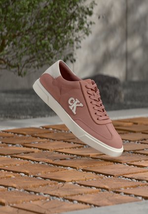 Roze en witte sneaker met "CK"-logo, zwevend boven een houten plankenvloer met kleine stenen, buiten met groen en een betonnen muur.