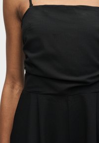 Γυναίκα που φοράει μαύρο φόρεμα χωρίς μανίκια με λεπτές τιράντες και εφαρμοστή μέση, εμφανίζεται από τον ώμο μέχρι το μέσο του μηρού σε απλό φόντο.