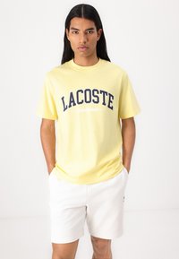 Gul bomull t-skjorte med marineblå grafikk "LACOSTE SPORTSWEAR" over brystet; kombinert med hvite shorts. Korte ermer og rund hals.