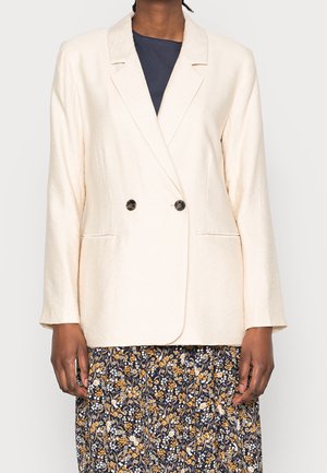 Person trägt ein cremefarbenes, zweireihiges Sakko über einem schwarzen Oberteil und einem marineblauen Rock mit gelbem und weißem Blumenmuster.