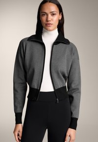 Szara i czarna kurtka na zamek z teksturowanym wzorem i wysokim kołnierzem, noszona na białym golfie, w połączeniu z czarnymi legginsami.