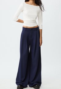 Femme portant un haut blanc à manches longues et court, un pantalon large bleu marine et des chaussures noires, debout devant un fond uni.