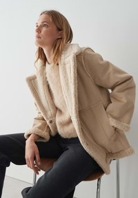 Manteau beige avec doublure et col doux et moelleux, doté d'une fermeture à boutons et de poches latérales. Porté sur un pull beige clair et un jean foncé.