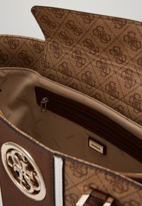 Brązowa torebka z monogramowym wzorem wewnątrz, z kieszenią na zamek błyskawiczny oraz dużym metalowym logo na przodzie.