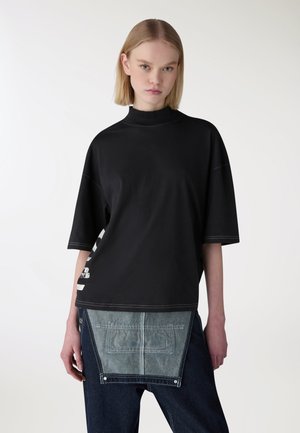 Giovane donna con capelli biondi corti che indossa una maglia nera larga con maniche a tre quarti e pantaloni in denim scuro con una grande toppa frontale.