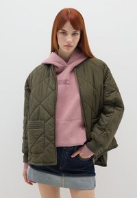 Giacca bomber trapuntata di colore verde oliva con polsini a costine, indossata sopra una felpa con cappuccio rosa. Il modello la abbina a una gonna di jeans.