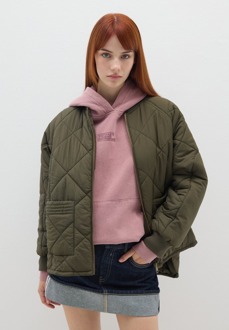 Giacca bomber trapuntata di colore verde oliva con polsini a costine, indossata sopra una felpa con cappuccio rosa. Il modello la abbina a una gonna di jeans.