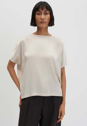Vrouw met kort donker haar, die een losse, korte mouw beige satijnen top en zwarte broek draagt tegen een effen lichte achtergrond.