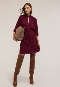 Burgunderfarbenes Kleid mit Schlüsselloch-Ausschnitt und gerafften Schultern, tailliert mit einem Bindegürtel. Braune kniehohe Stiefel mit Absatz und eine hellbraune Handtasche.