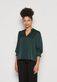 Blouse en satin vert foncé avec un décolleté en V, des manches froncées et une coupe ample. Assortie à un pantalon noir, avec une texture lisse et brillante.