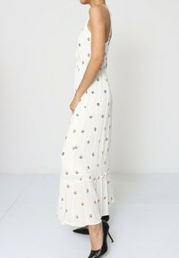 Robe maxi blanche avec des embellissements floraux dorés, bretelles ajustables et ourlet à volants. Portée avec des talons hauts noirs en tissu texturé.