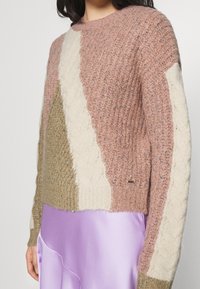 Strickpullover mit einem Patchwork-Design in Pink, Beige und Creme, mit strukturiertem Rippstrick und Zopfmusterakzenten. Getragen über einem lavendelfarbenen Rock.