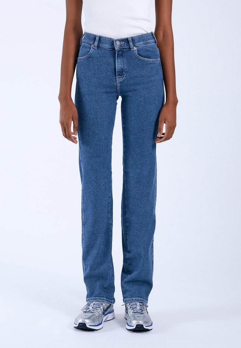 Person iført blå straight-leg jeans, en hvid ærmeløs top og sølvfarvede sneakers, stående på en hvid baggrund.
