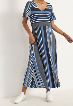 Vrouw die een lange V-halsjurk met korte mouwen draagt, met blauw, zwart en wit gestreept patroon, en witte sneakers, staand tegen een effen achtergrond.