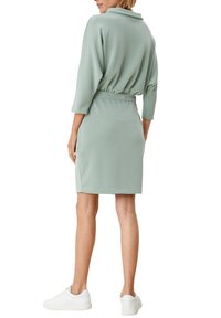 Vihreä sametti-mekko, jossa on kuminauhavyötärö, 3/4-hihat ja kaulus, yhdistettynä valkoisiin lenkkareihin. Sileä pinta, keskikokoinen muotoilu.