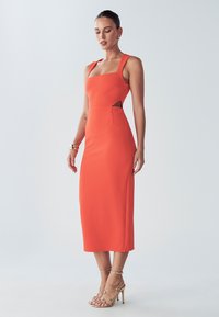 Orange tætsiddende kjole med firkantet halsudskæring, udskæringsdetalje i siden og længde til midt på læggen. Kompletteret med gyldne hæle med stropper.