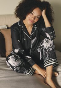 Fekete selyem pizsamaszett szürke és fehér árnyalatú virágmintával. A nadrágon csíkos mandzsetta található. A modell egy ágyon ül.