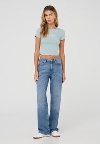 T-shirt corto azzurro chiaro con stampa a logo, abbinato a jeans blu a vita alta e gamba larga e sneakers bianche su uno sfondo semplice.