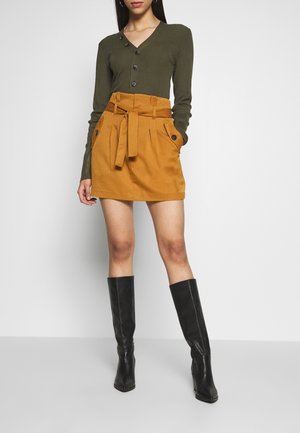 Kobieta ubrana w oliwkowo-zielony sweter na guziki, musztardowo-żółtą mini spódnicę z paskiem oraz czarne, wysokie kozakii na obcasie, stojąca na białym tle.