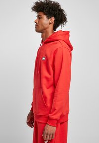 Red hooded sweatshirt made of cotton blend fabric, featuring a kangaroo pocket and a small logo patch on the chest. Soft texture.