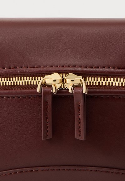 Κοντινό πλάνο δύο χρυσών φερμουάρ σε κλειστή καφέ δερμάτινη τσάντα με λεπτομερείς ραφές στις άκρες.