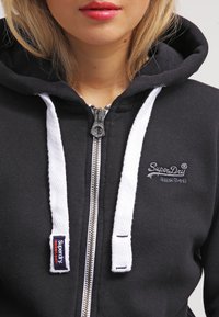 Sudadera negra con cremallera y cordones blancos, presenta una cremallera plateada y un logotipo bordado. Tela suave con un diseño informal.