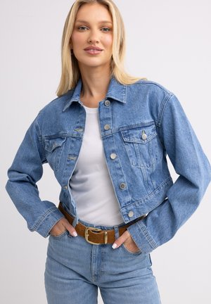 Chaqueta de mezclilla azul clara con un diseño recortado, parte delantera con botones, dos bolsillos en el pecho y textura desgastada. Llevada sobre una camiseta blanca. Cinturón marrón como acento.