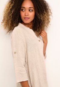 Beige linen button-up dress with three-quarter sleeves, featuring a collar and a button accent on rolled sleeves. Textured fabric visible.